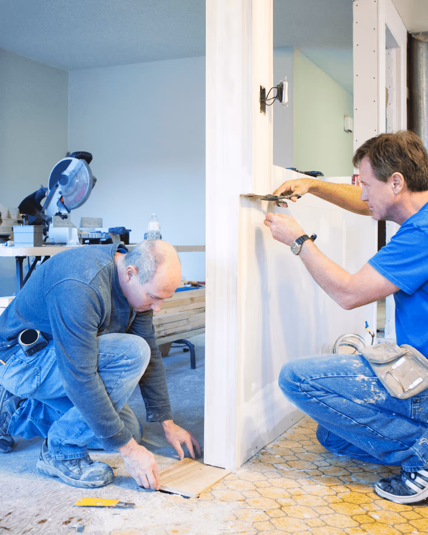 Zwei Handwerker bei der Verlegung von Holzboden und dem Spachteln einer Wand in einem Innenraum.