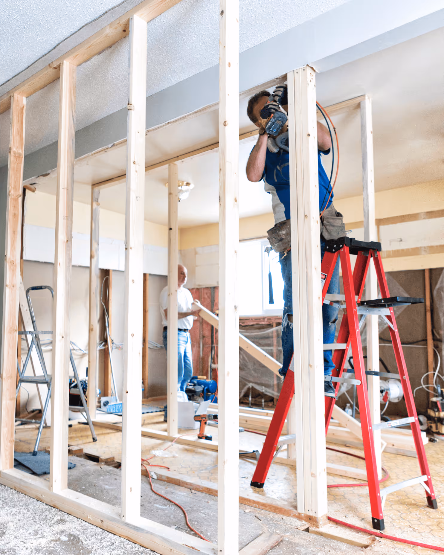Zwei Handwerker bauen eine Innenwand in einem Raum mit Holzrahmen; einer steht auf einer roten Leiter und arbeitet am oberen Teil, der andere hält ein Holzbrett.