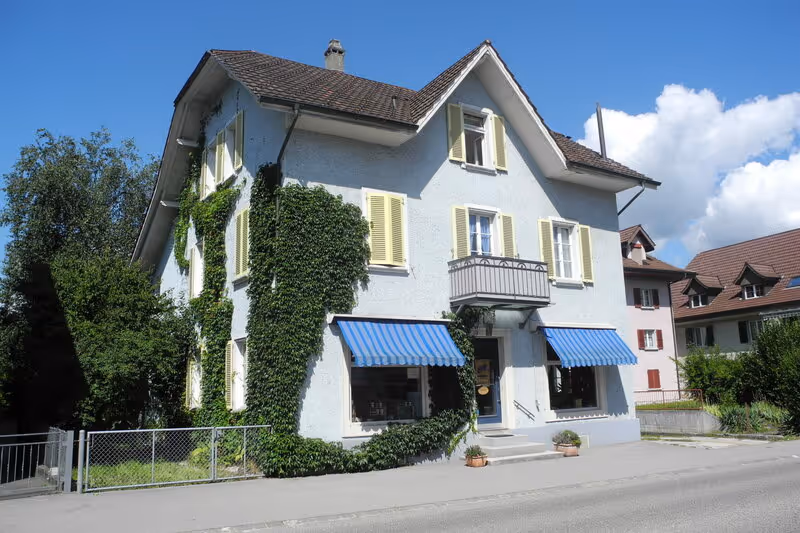 Weisses, mehrstöckiges Haus mit gelben Fensterläden und blauen Markisen, teilweise begrünt, an einer ruhigen Strasse bei klarem Himmel.