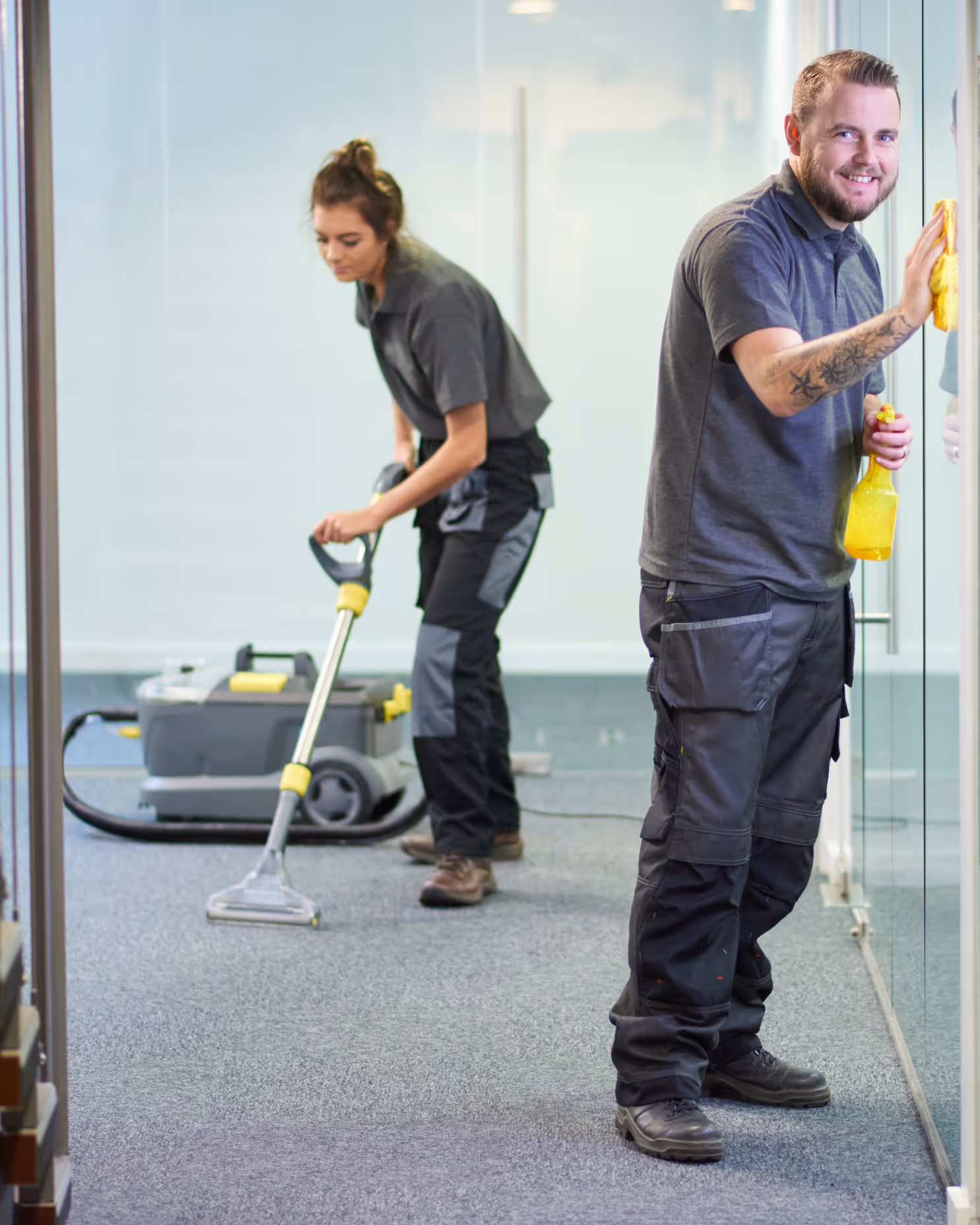 Zwei Reinigungskräfte in dunkler Arbeitskleidung reinigen einen Büroraum, eine wäscht ein Glas und die andere saugt den Teppichboden.