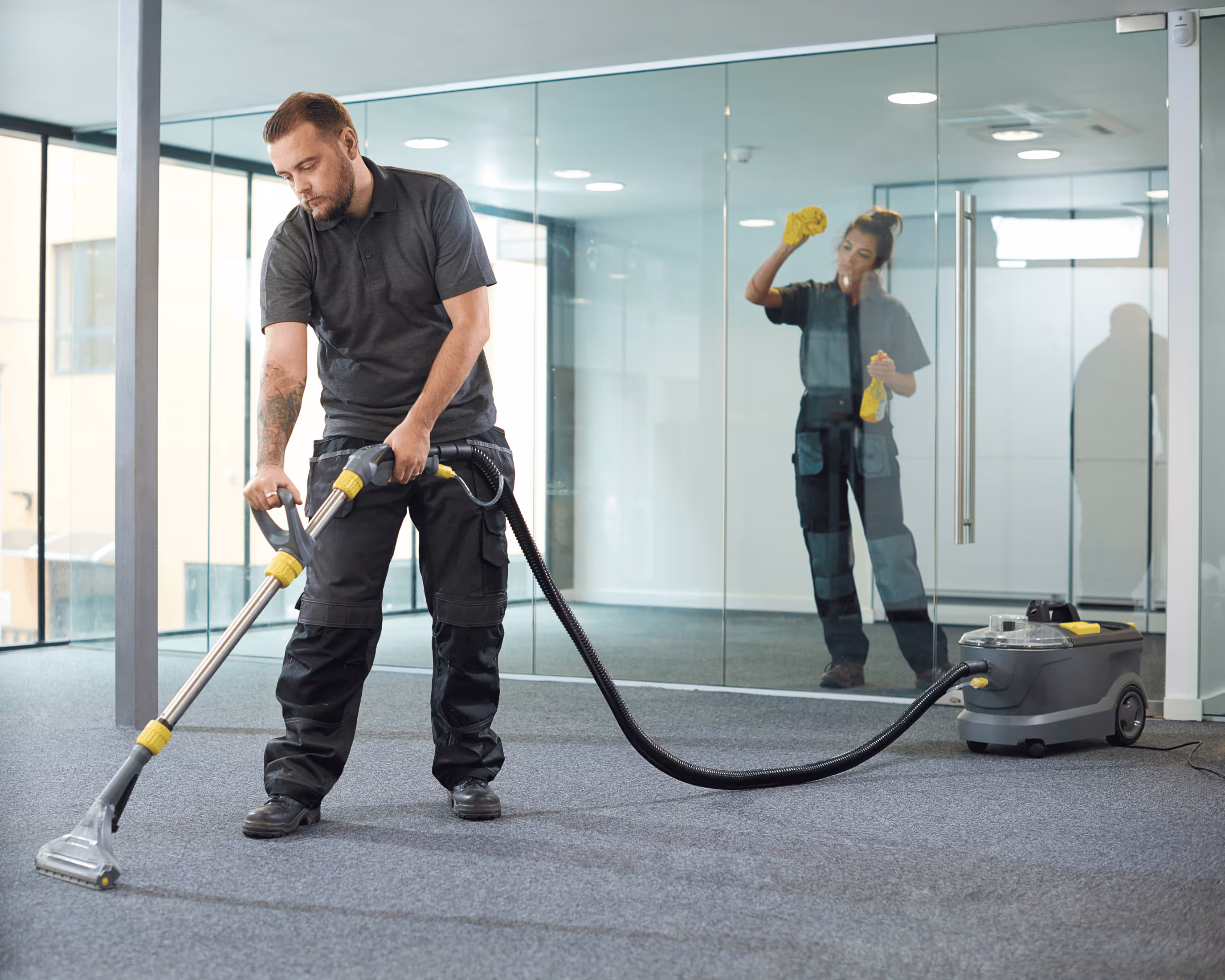 Ein Mann reinigt mit einem Sauger den Teppichboden in einem Büro, während eine Frau im Hintergrund eine Glaswand putzt.