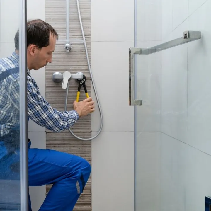 Man in blue overalls repairing a shower faucet with a wrench inside a modern bathroom.