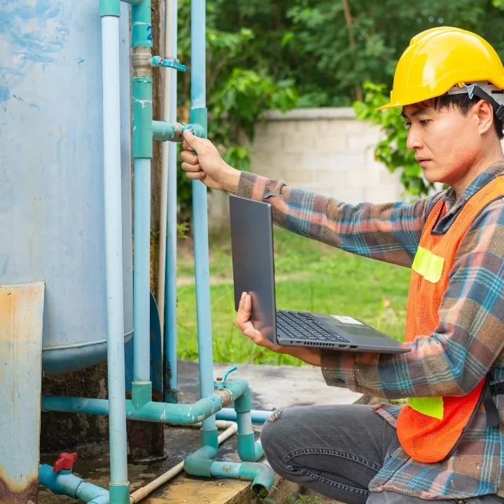 Worker in a yellow hard hat and orange safety vest holding a laptop and adjusting a valve on outdoor plumbing pipes.