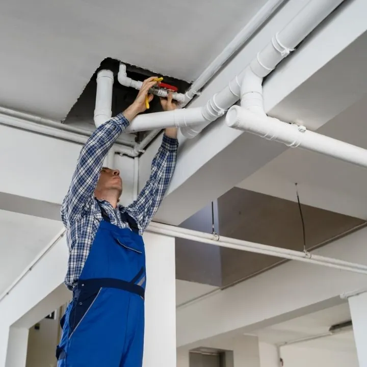 Plumber in blue overalls and plaid shirt repairing white ceiling pipes in a building.
