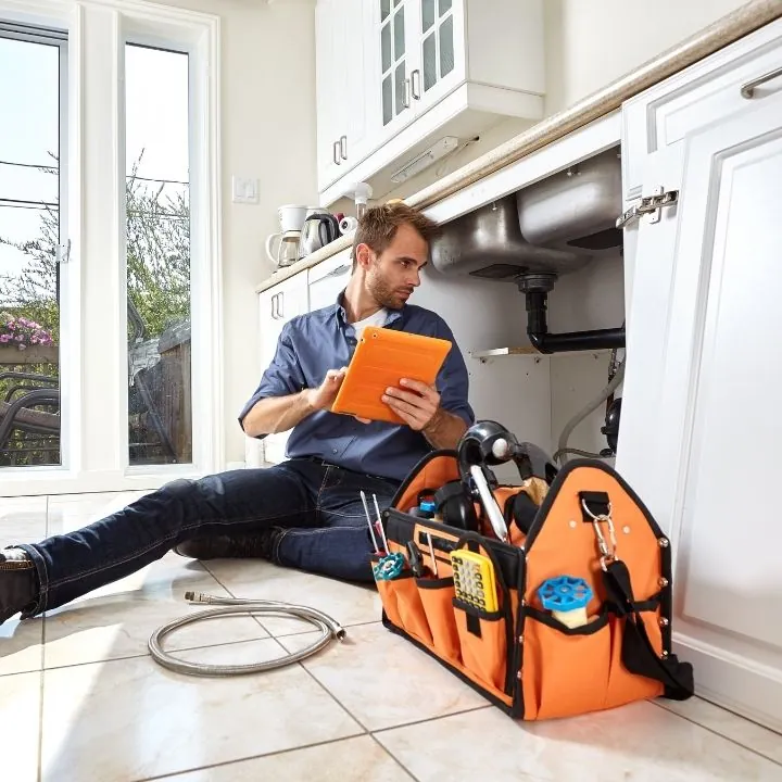 Plumber sitting on kitchen floor examining plumbing under sink while holding an orange tablet, next to an orange tool bag and plumbing tools.
