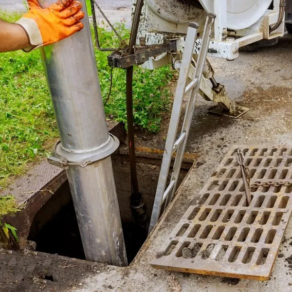 Worker wearing orange glove lowering a metal pipe into an open sewer manhole with a ladder and a partially removed grate on the side of a street.