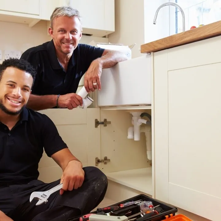Two plumbers smiling and posing near a kitchen sink with an open cabinet showing pipes and a toolbox on the floor.