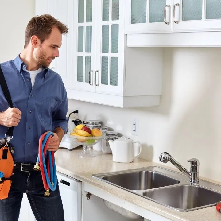 Plumber holding red and blue hoses standing in a kitchen next to a double sink with white cabinets and countertop.