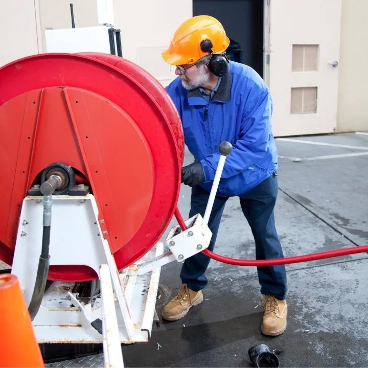 Worker in safety helmet and blue jacket handling a large red hose reel outdoors.