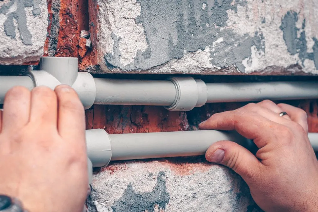 Hands holding and installing gray PVC pipes against a rough brick and plaster wall.