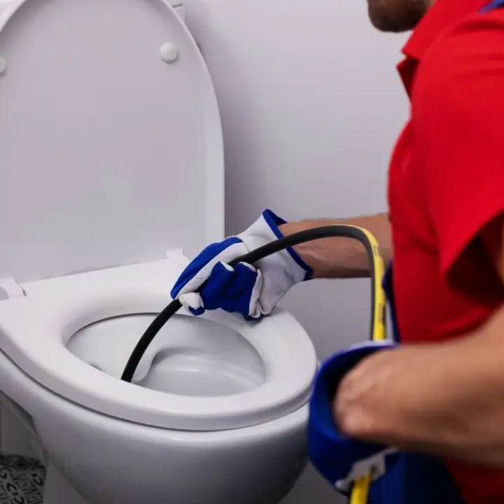 Person wearing gloves and a red shirt using a plumbing snake to unclog a white toilet.