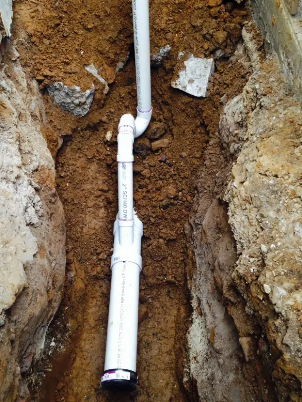 White PVC drainage pipes installed in a narrow dirt trench with soil and rocks surrounding the pipes.