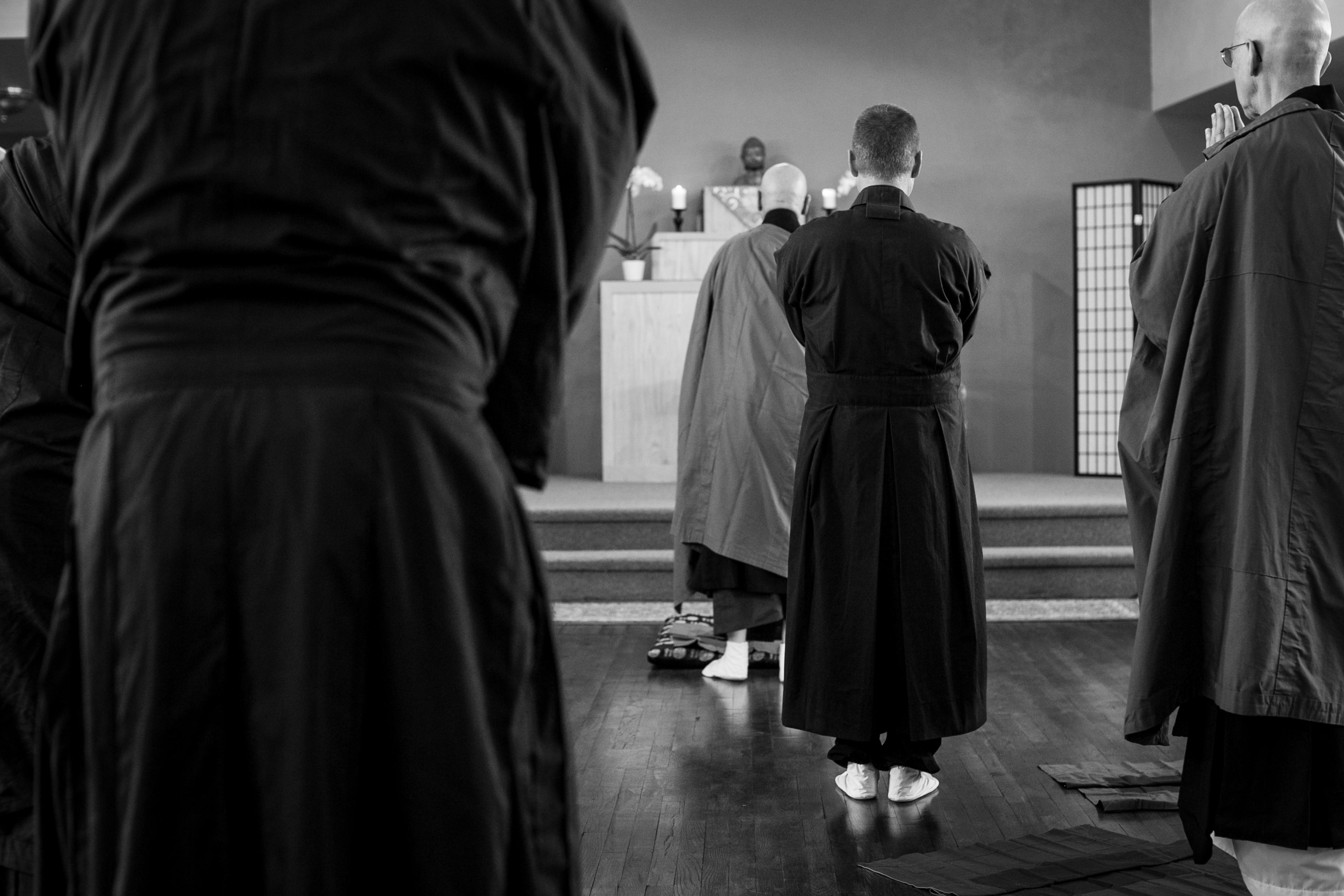 Monks in robes standing in meditation facing an altar with a Buddha statue and candles.