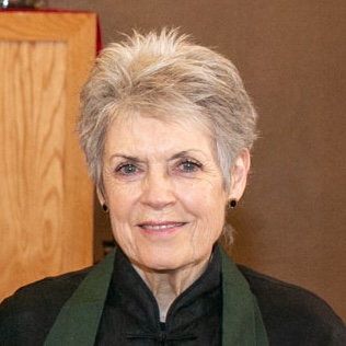 Smiling woman with short hair wearing a black top and dark green scarf against a brown background.