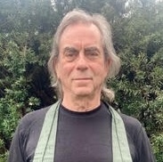 Man wearing a black shirt and light green apron standing outdoors with greenery in the background.