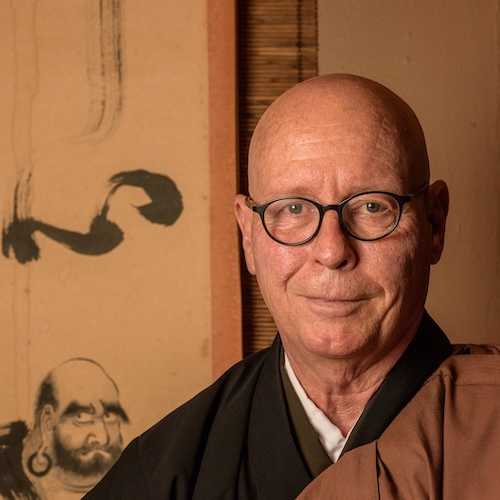 Man with glasses wearing traditional Buddhist robes standing in front of a scroll with an ink painting on the wall.