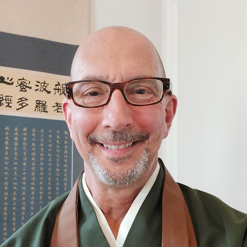Man with glasses wearing a brown and green traditional robe, standing indoors with framed calligraphy in the background.