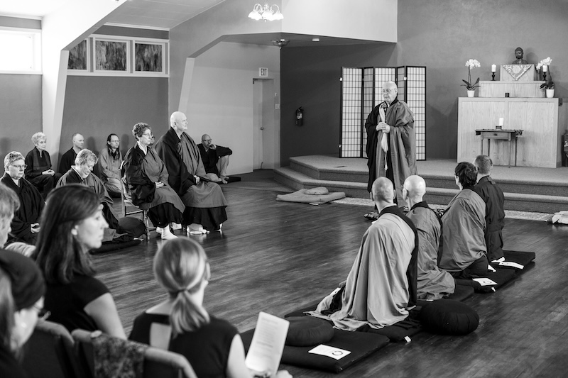 Monk standing and speaking in a meditation hall with a seated audience, some wearing traditional robes.