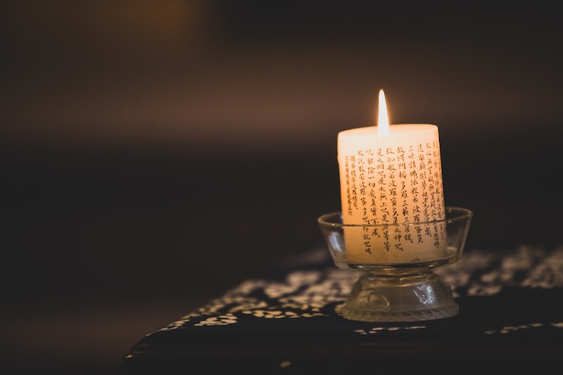Lit candle with Asian script characters on its surface placed in a glass holder on a patterned cloth.