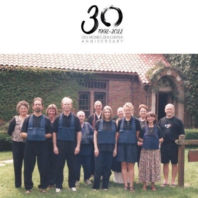 Group of eleven people standing on grass in front of a brick building with ivy and a tiled roof, some wearing blue cloth aprons.