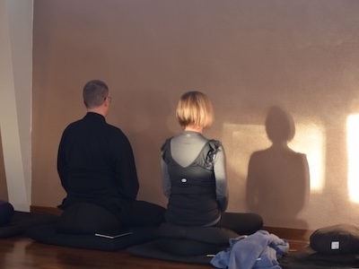 Two people sitting cross-legged on cushions meditating facing a wall with sunlight and shadows.