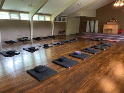 Room with wooden floor arranged with black meditation cushions and mats in rows facing a small elevated platform with a wooden altar.