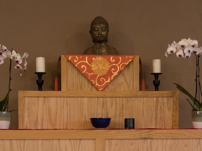 Wooden altar with a Buddha statue, flanked by two white orchids in pots and two white candles on black holders.