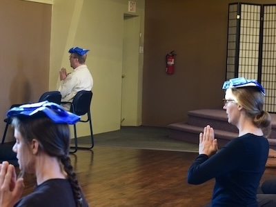 Three people sitting cross-legged on the floor and a chair in a meditation pose with blue cloths on their heads in a calm studio room.