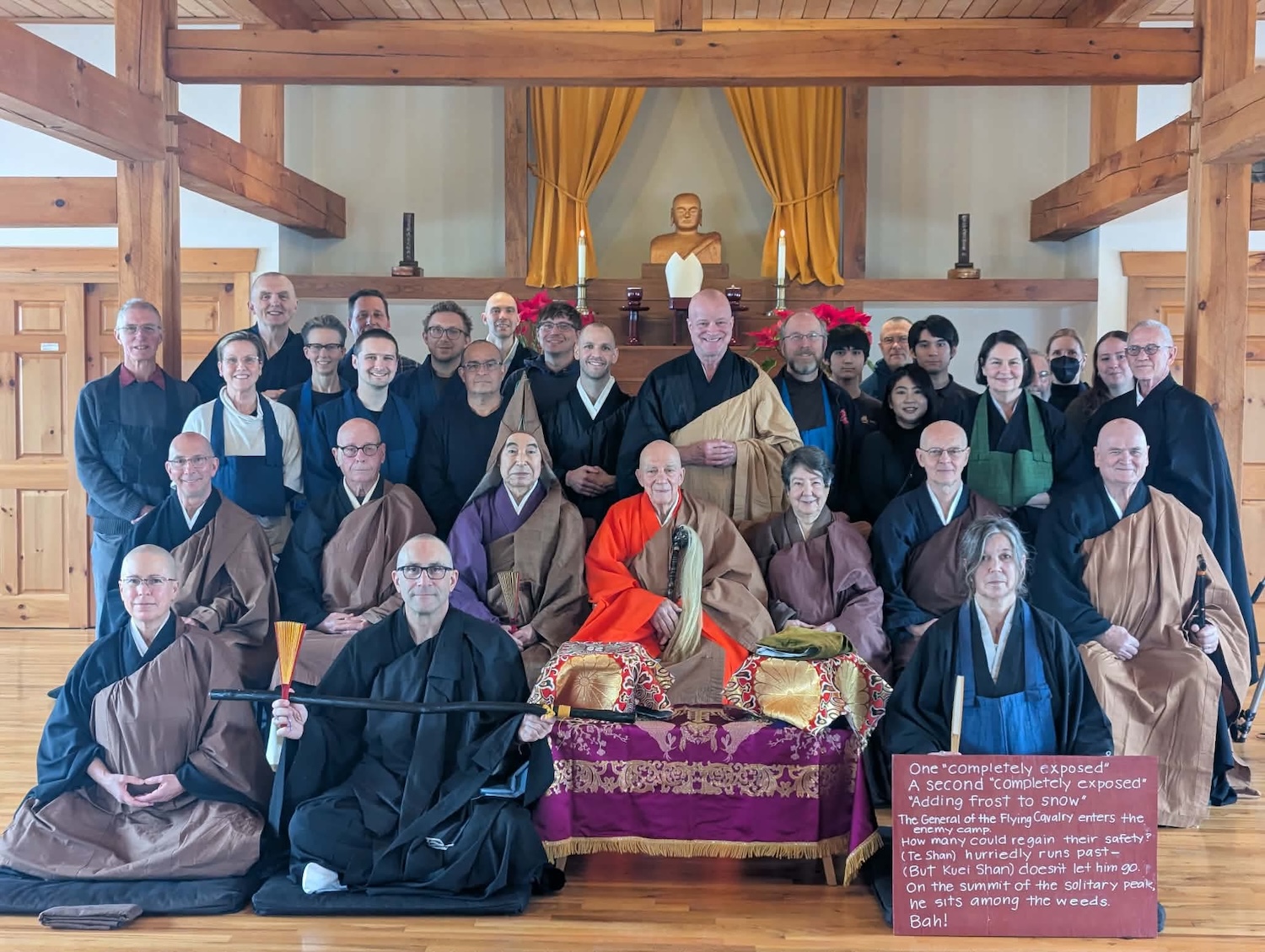 Group of monks and practitioners in traditional robes posing in a wooden temple with a Buddha statue and candles in the background.