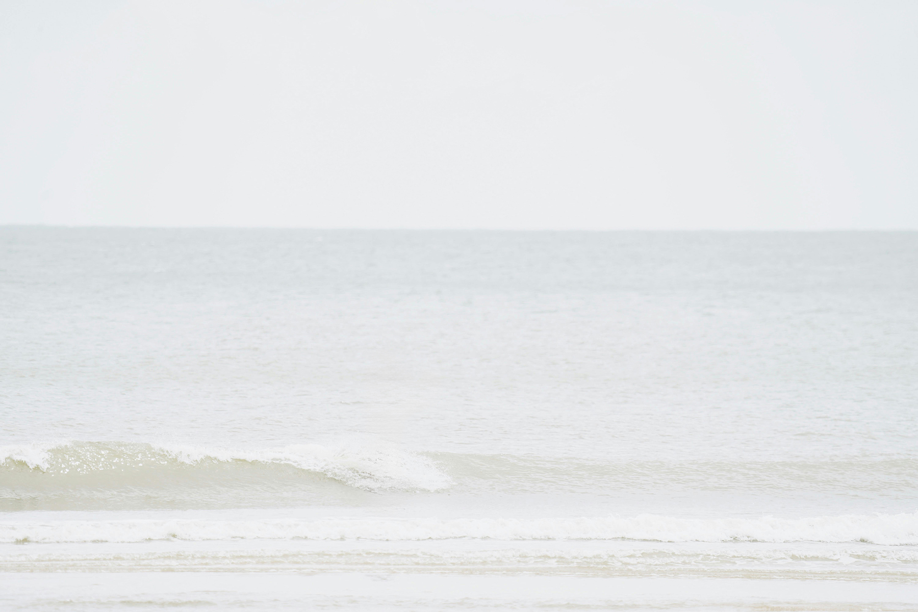 Gentle ocean waves rolling onto a calm sandy beach under a pale, overcast sky.