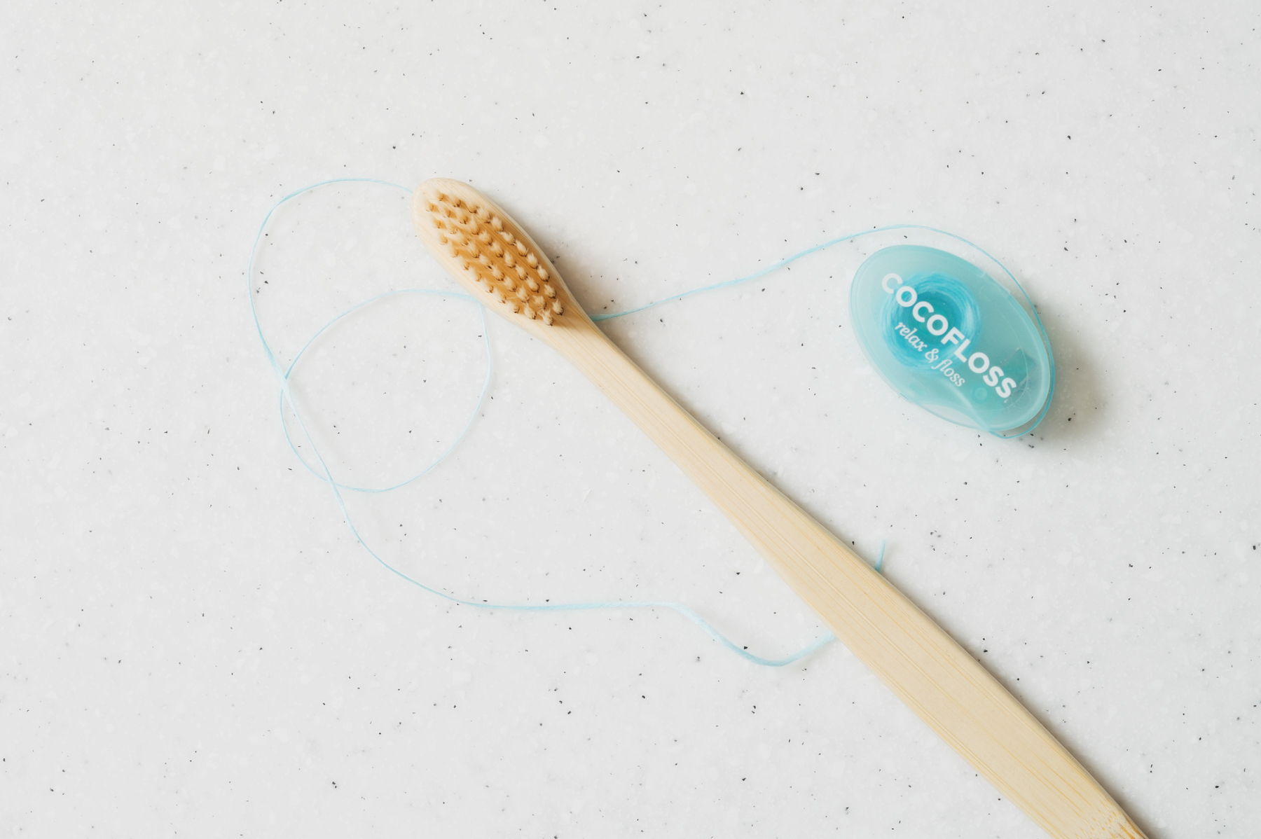Bamboo toothbrush with beige bristles next to blue dental floss container labeled 'COCOFLOSS' on a white speckled surface.