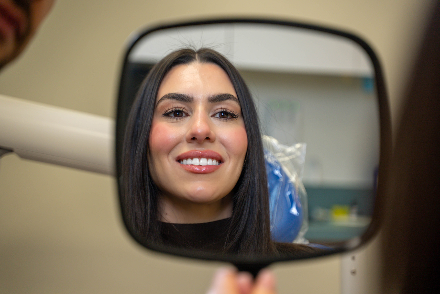 Woman with long dark hair smiling and looking at her reflection in a handheld mirror in a dental office.