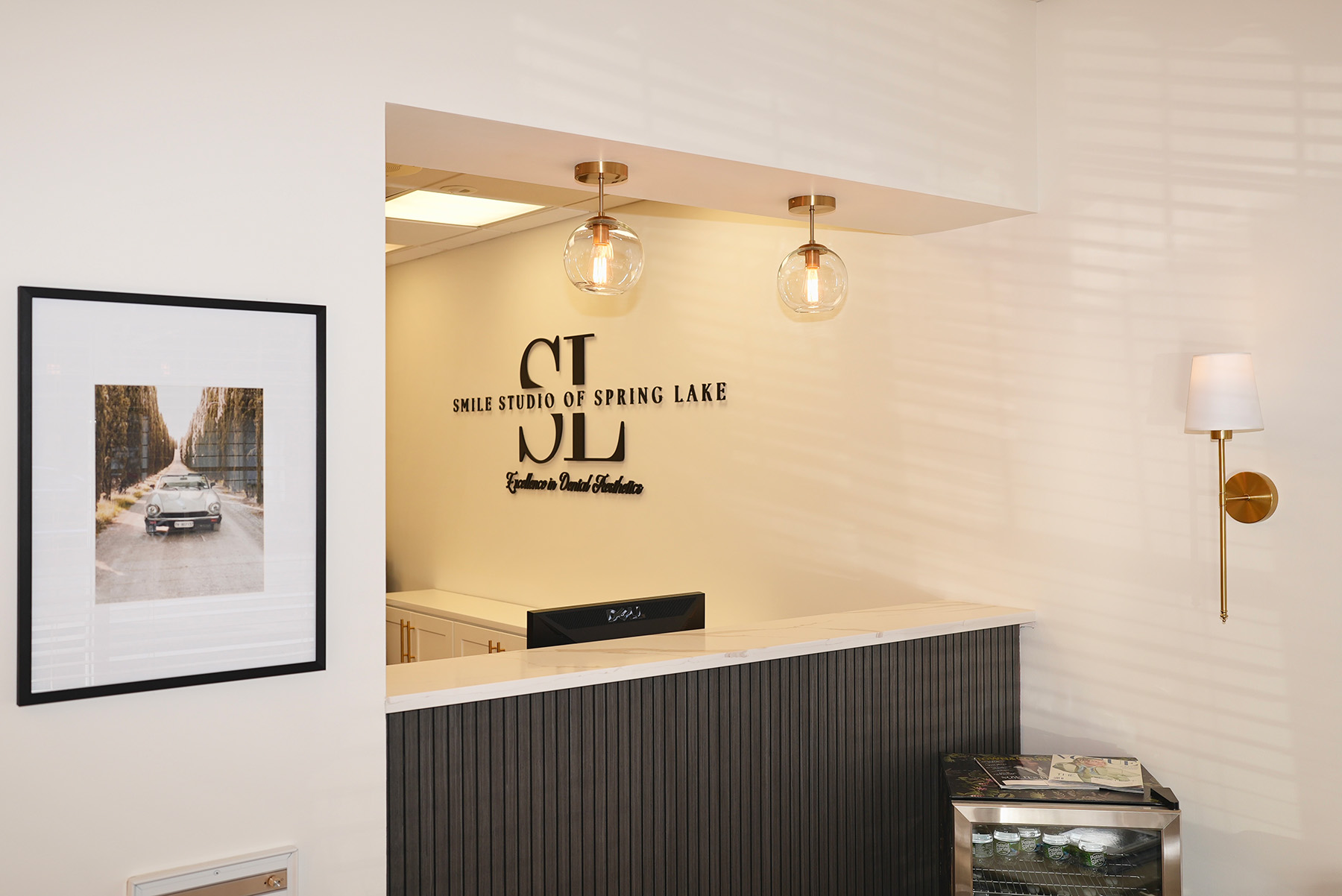 Reception area of Smile Studio of Spring Lake with a marble countertop, two hanging glass pendant lights, framed car photo on wall, and a small fridge with magazines on top.