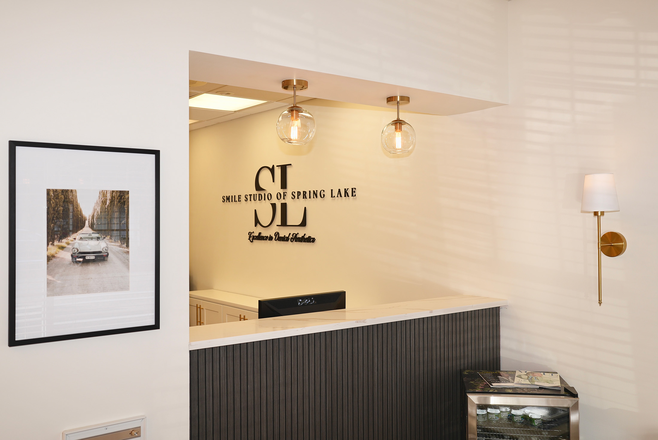Reception desk at Smile Studio of Spring Lake with logo on wall, two hanging globe lights, framed car photo, and small beverage fridge.