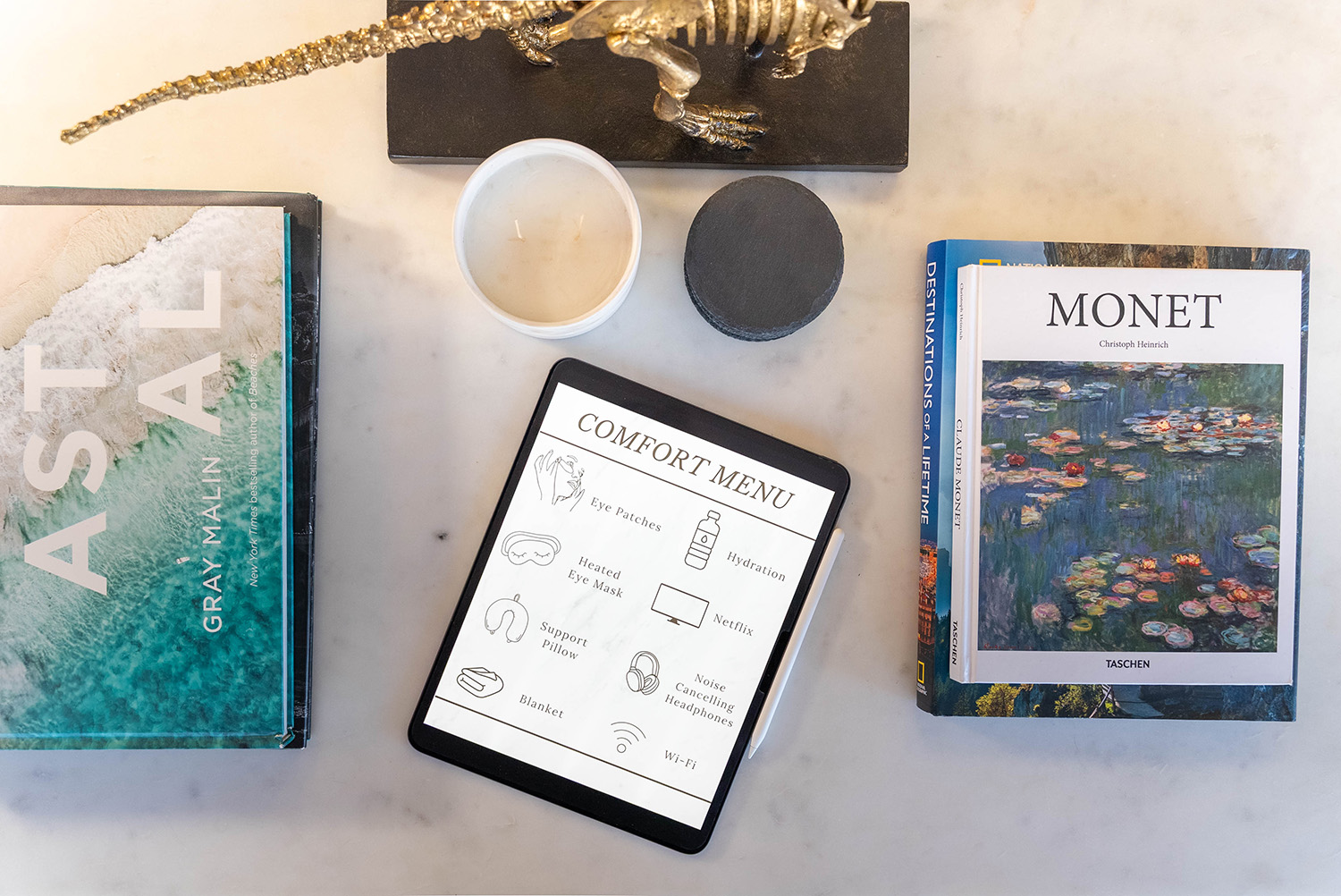 Top view of a tablet displaying a comfort menu with items like eye patches, hydration, Netflix, and Wi-Fi, surrounded by a candle, a dinosaur figurine, and art books titled Astoral and Monet.