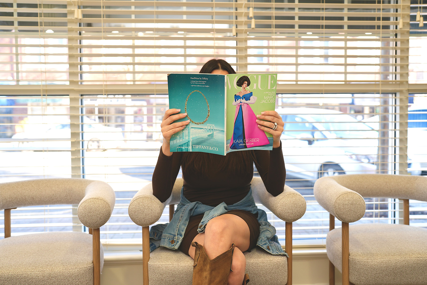 Person sitting on a beige armchair reading a Vogue magazine with a Tiffany & Co. advertisement, with window blinds and parked cars visible behind.