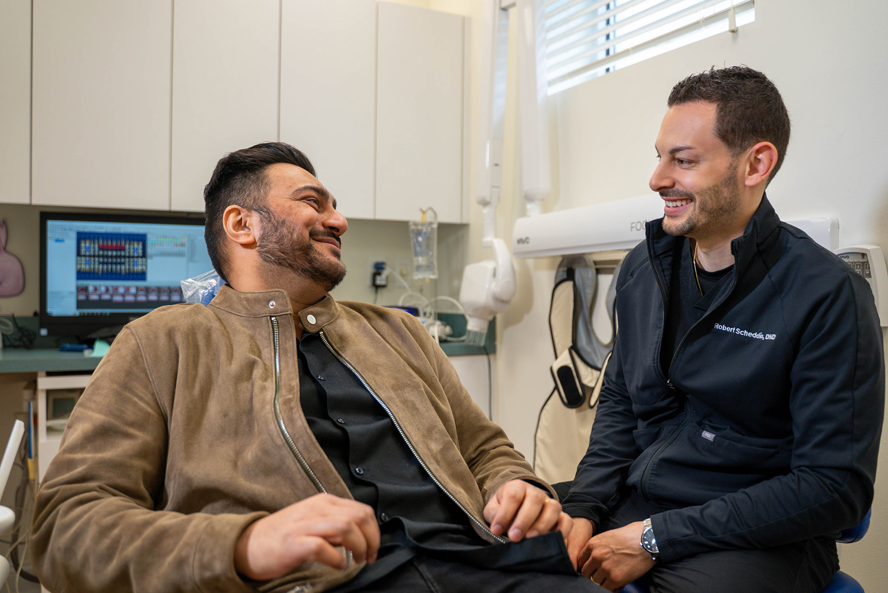 Dentist in black jacket smiling and talking with a relaxed male patient in a dental clinic.