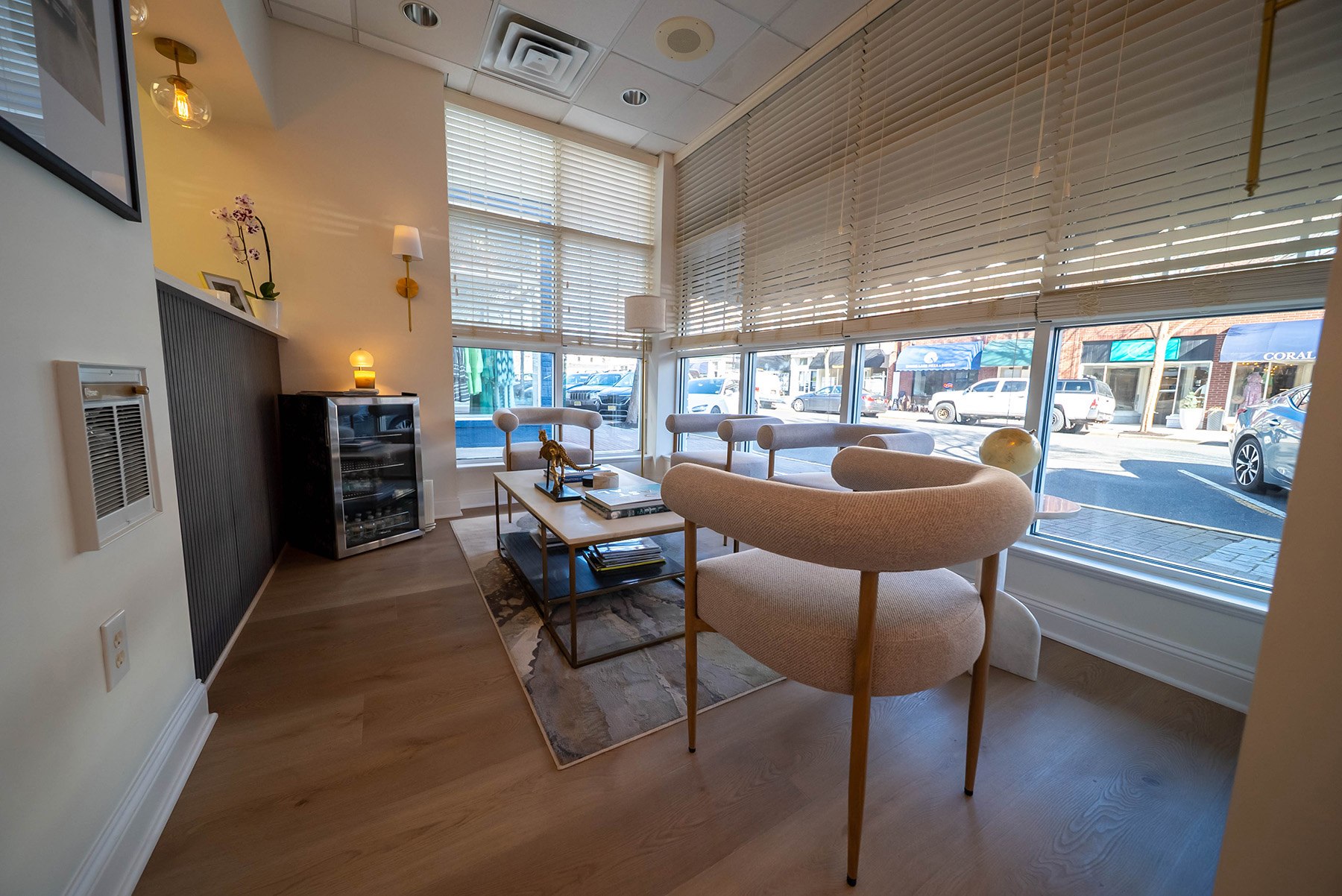 Modern waiting area with curved beige chairs, coffee table with books and decor, large windows with blinds overlooking parked cars outside.
