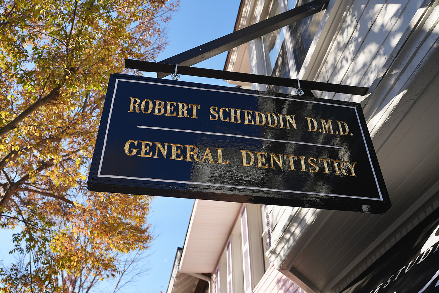 Black hanging sign with gold lettering reading 'Robert Scheddin D.M.D. General Dentistry' outside a building with autumn trees and blue sky in the background.