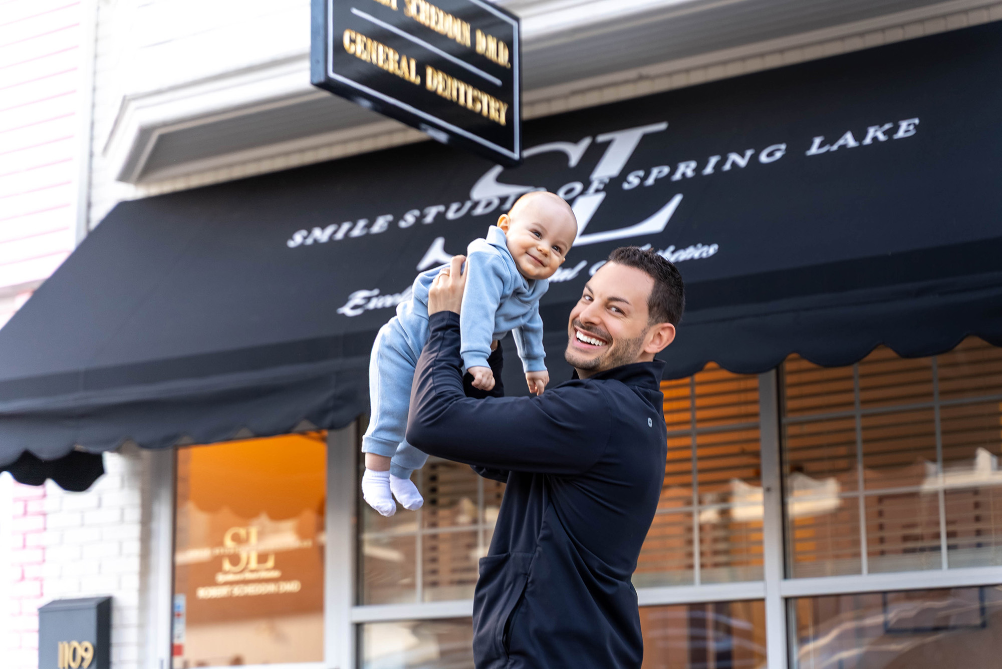 Smiling man in a dark jacket holding a smiling baby in a blue outfit outside a building with a black awning that reads Smile Studio of Spring Lake.