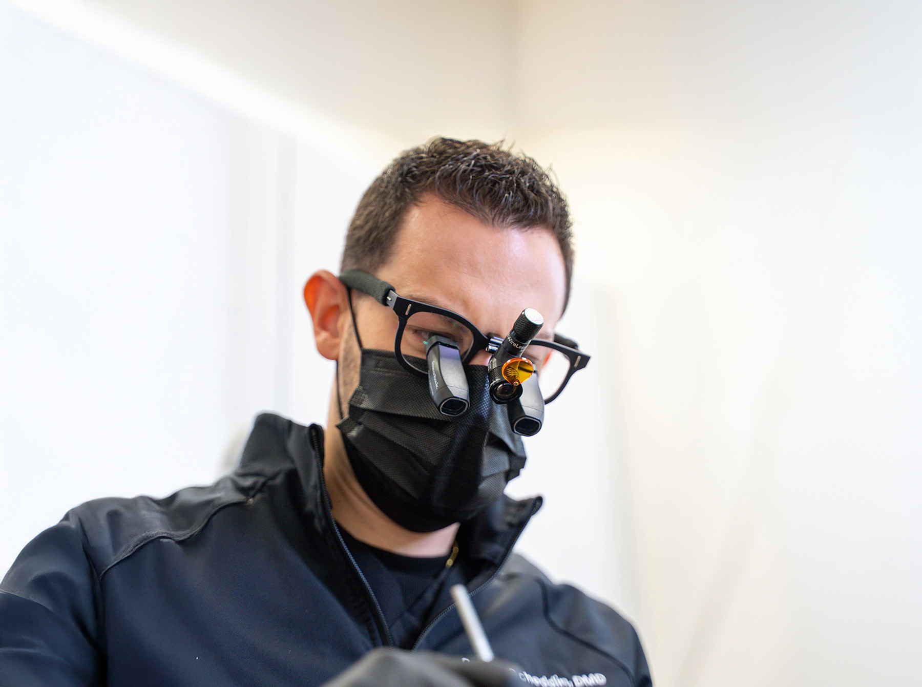 Dentist wearing magnifying loupes and a black face mask, focusing on a dental procedure.