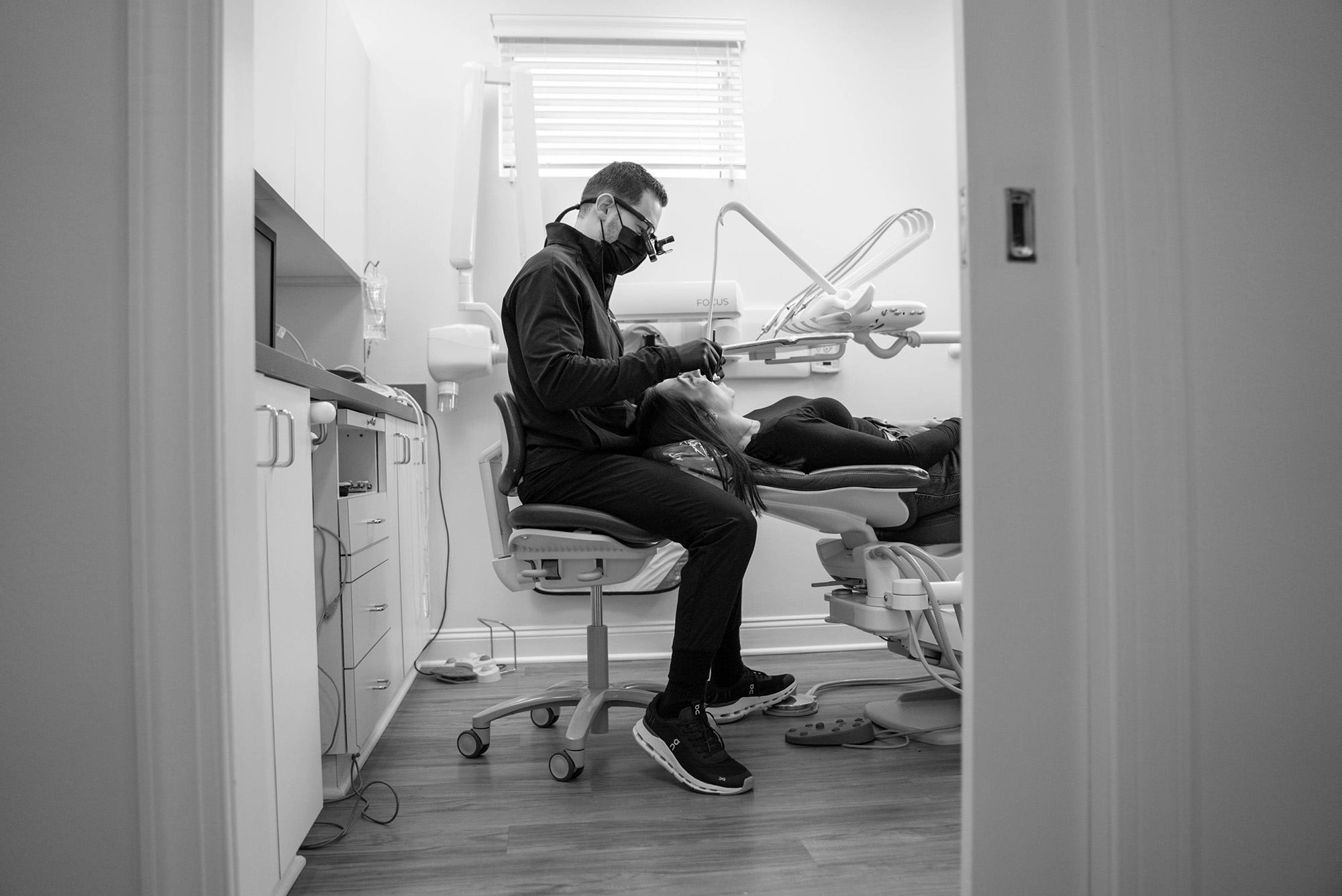 Dentist wearing magnifying glasses and mask treating a patient lying back in a dental chair in a clinic room.