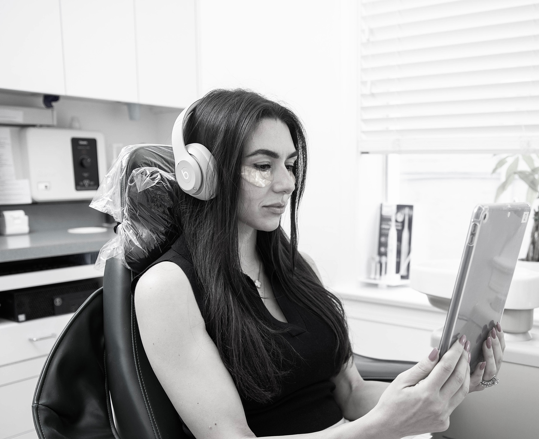 Woman with under-eye patches wearing headphones and reading a tablet while sitting in a dentist's chair.