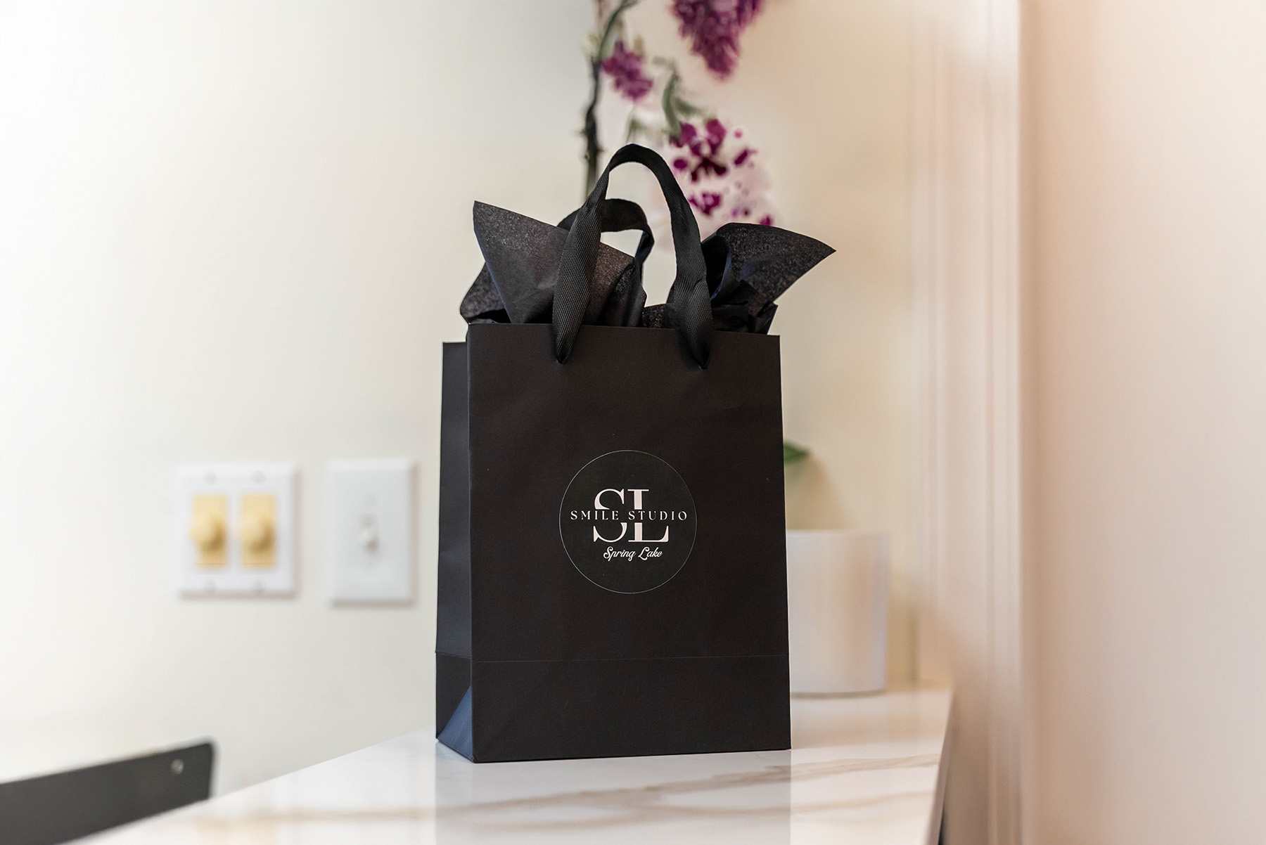 Black gift bag with black tissue paper and a circular Smile Studio Spring Lake logo, placed on a white marble countertop with a blurred background.