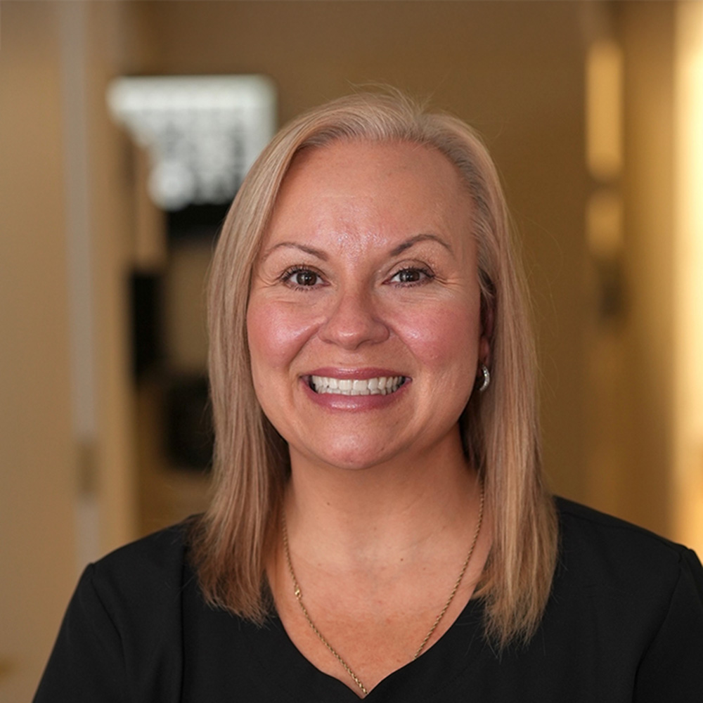 Smiling woman with straight blonde hair wearing a black top and gold necklace, indoors with blurred background.