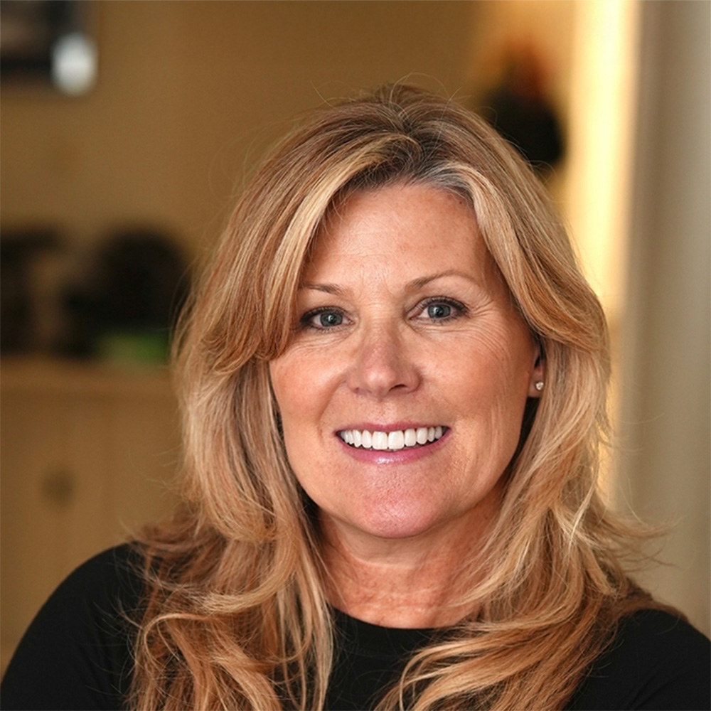 Smiling middle-aged woman with long blonde hair wearing a black top indoors.
