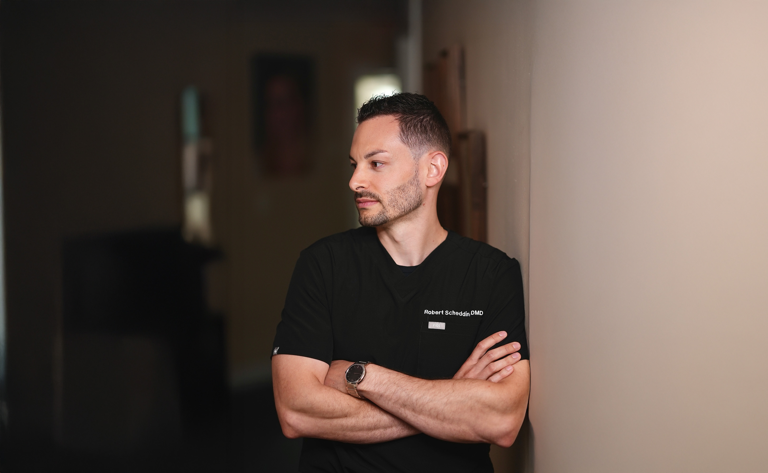 Man with short dark hair and beard wearing black scrubs with name tag Robert Scheddin, DMD, standing with arms crossed against a beige wall.