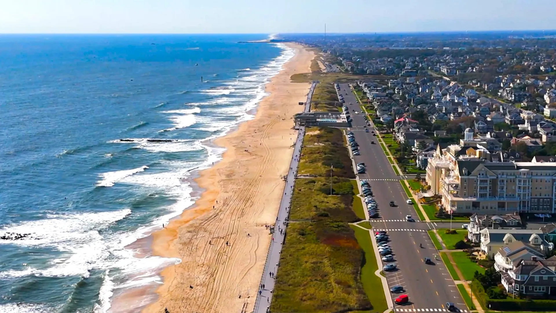 Aerial view of a coastline featuring a sandy beach, ocean waves, a boardwalk, a road with parked cars, and residential buildings.