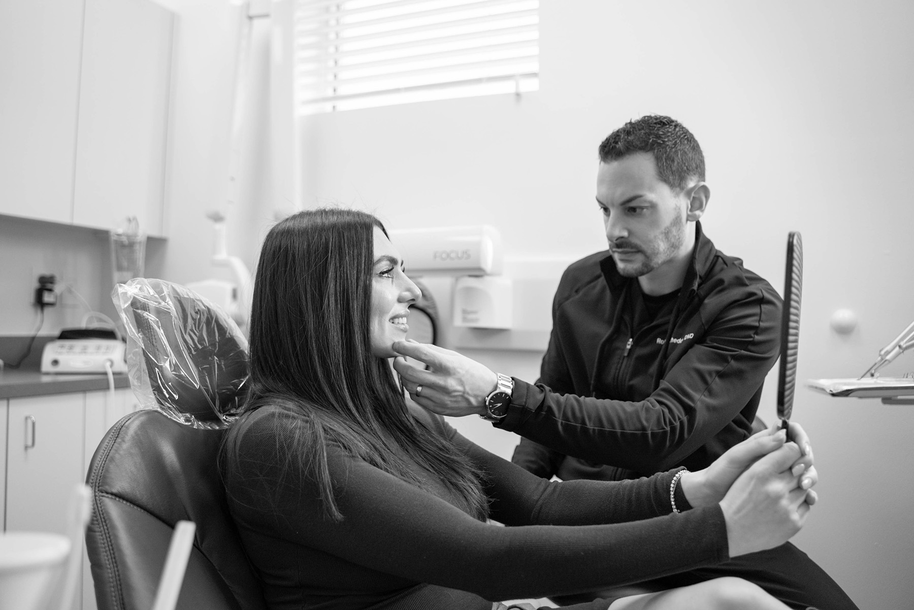 Dentist examining a female patient’s smile as she holds a mirror in a dental office.