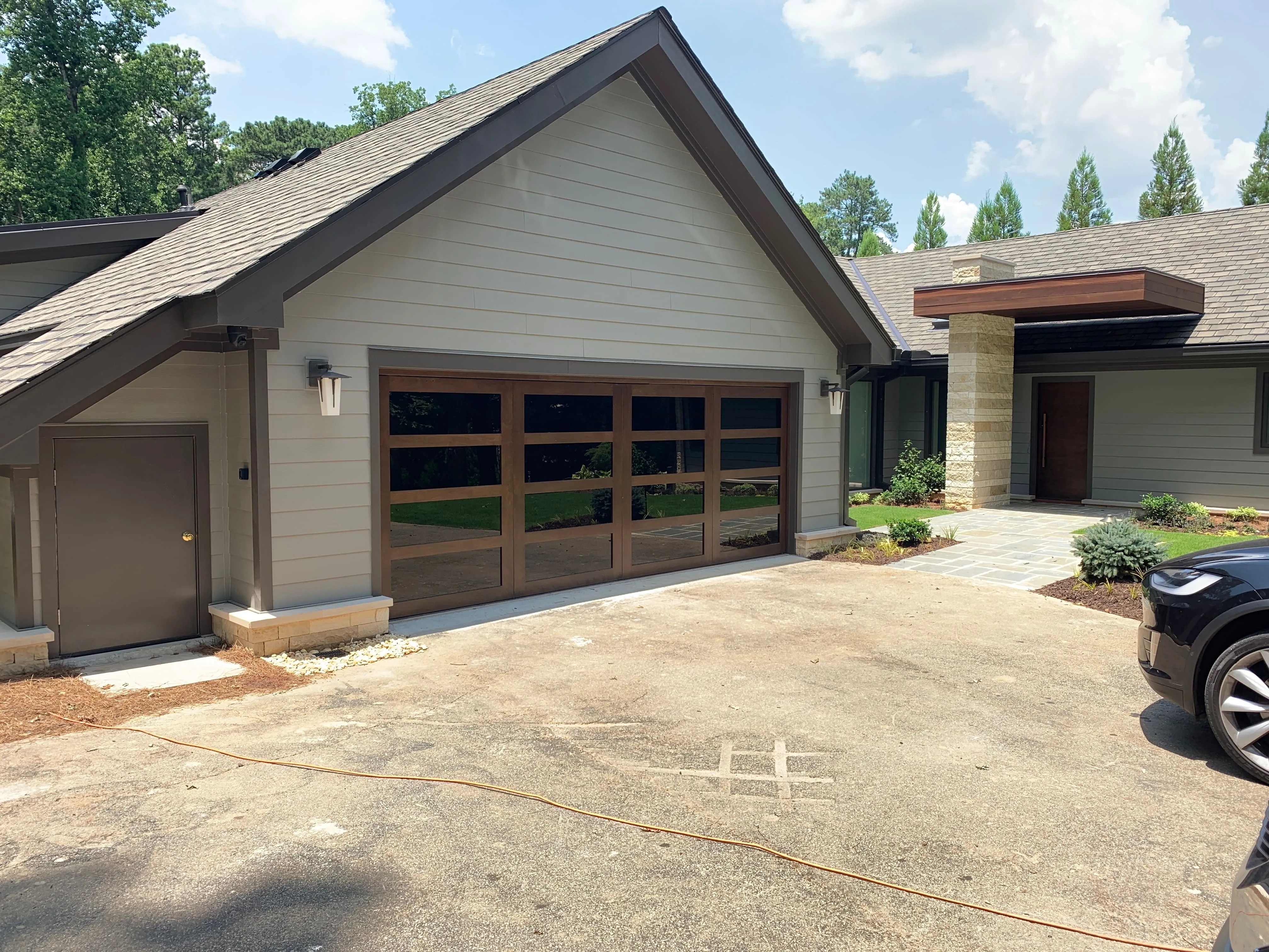 Modern house exterior with a large wooden-framed glass garage door, gray siding, stone chimney, and driveway with a black car partially visible.